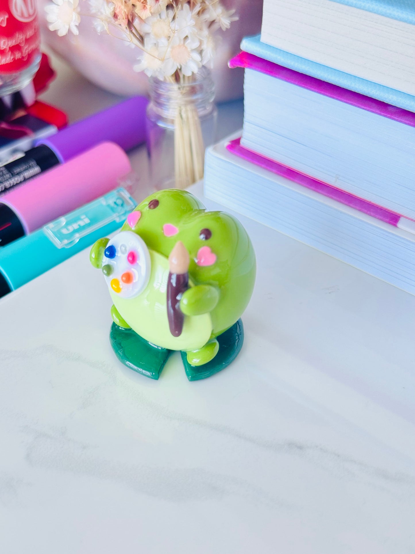 Green frog figurine holding a pencil on a desk with stationery items in the background.