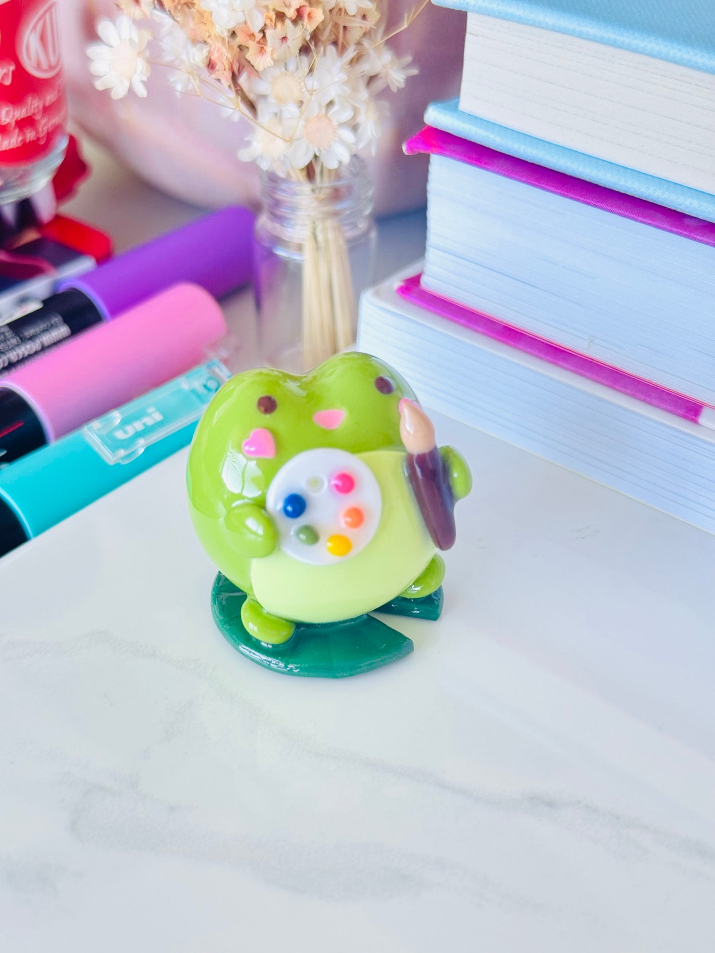 Colorful frog figurine with a pencil on a desk with stationery items.