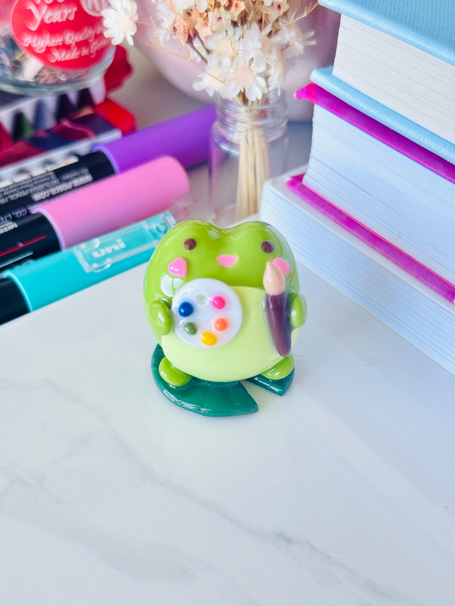 Colorful frog figure on a desk with markers and books in the background