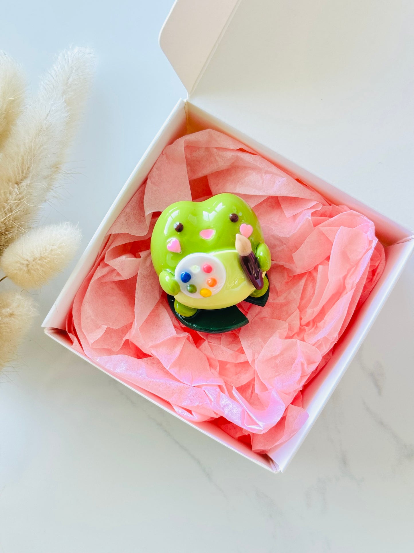 Green frog figurine with colorful paintbrushes in a box with pink tissue paper on a light background