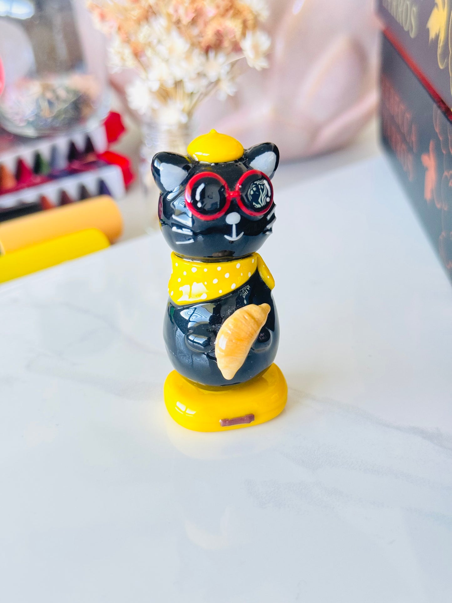 Black cat figurine with red glasses and yellow hat on a white surface