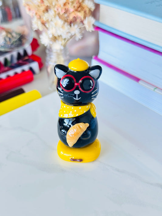 Black cat figurine with yellow hat and red glasses on a white surface