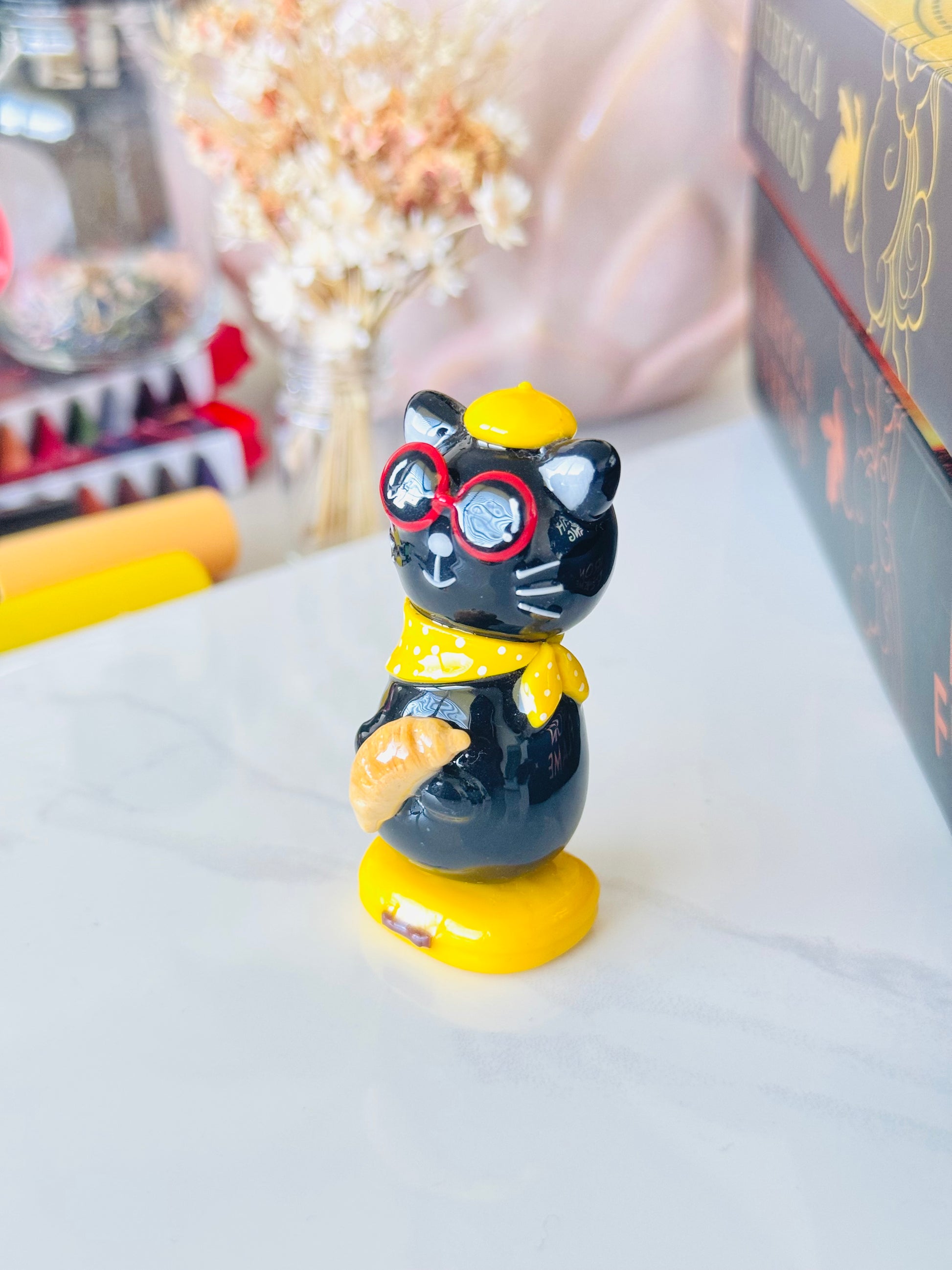 Black cat figurine with red glasses and a yellow bow tie on a white surface.