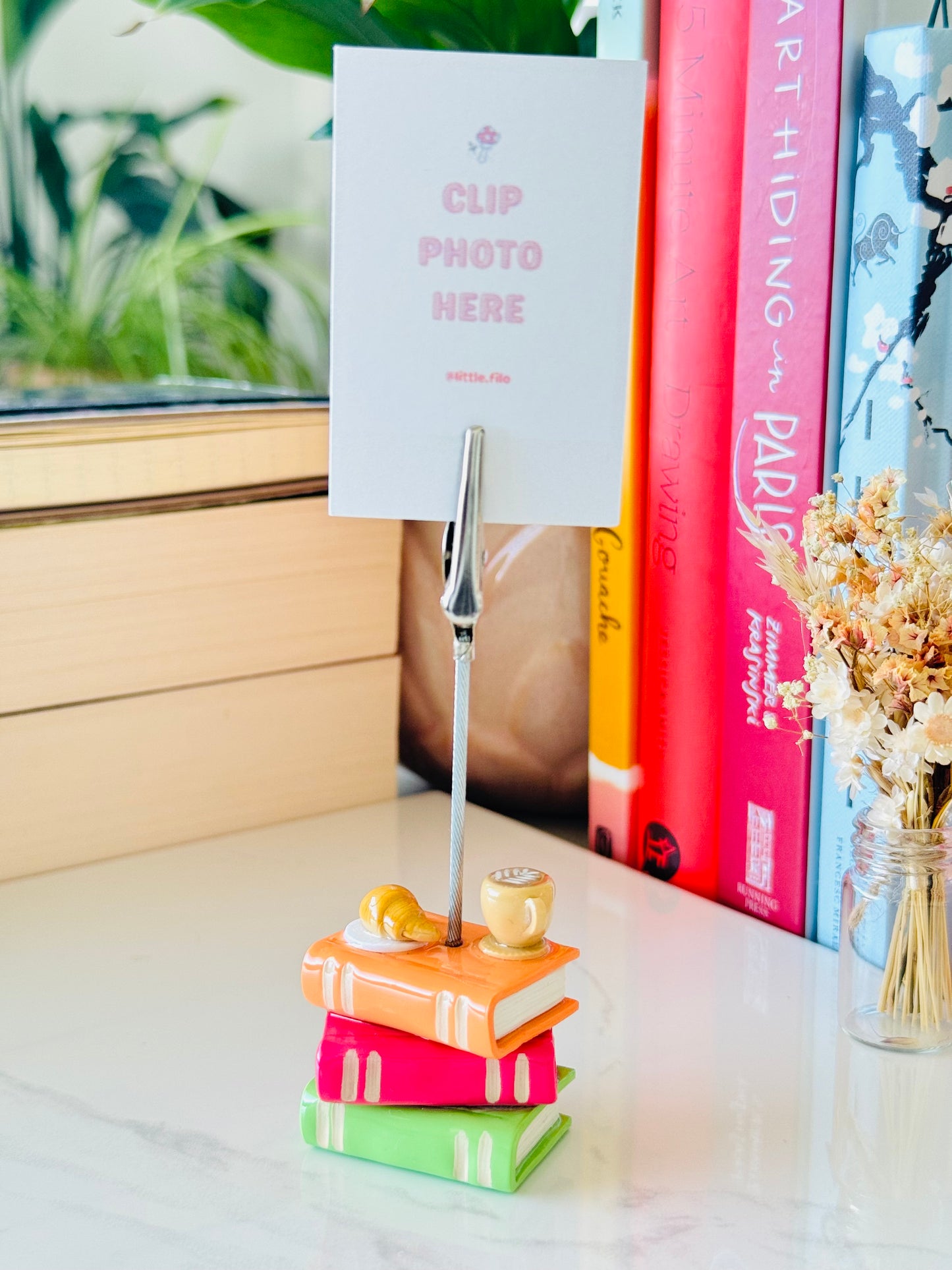 Notebook clip holder with a photo card on top, placed on a stack of books with a window and plant in the background.