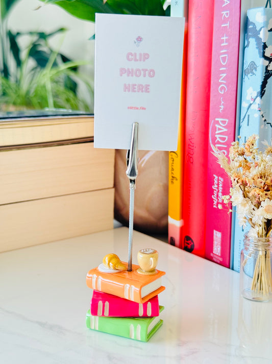 Notebook clip holder with a photo card on top, placed on a stack of books with a window and plant in the background.