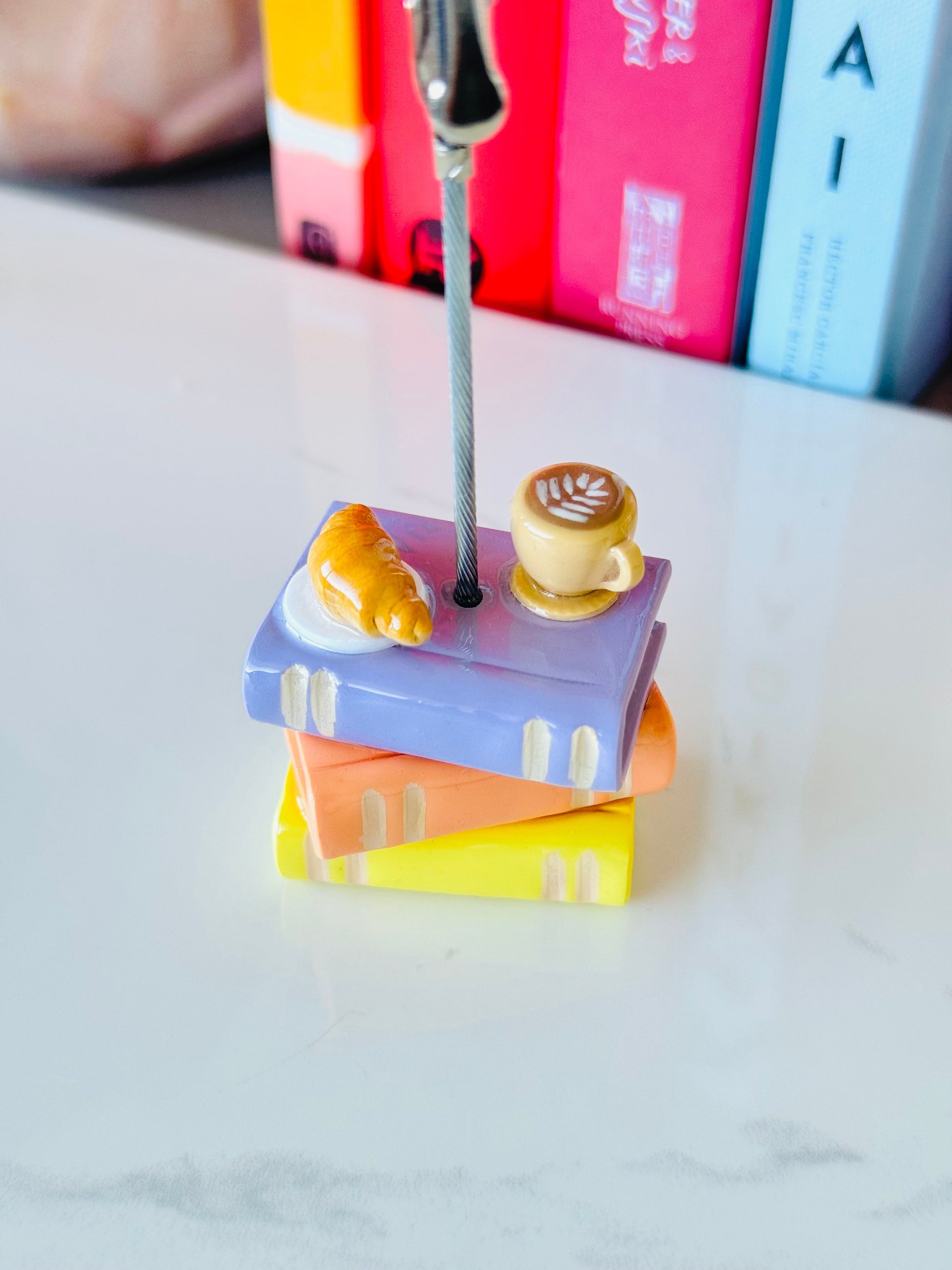 Colorful miniature books with a croissant and coffee cup on top, held together by a spoon.