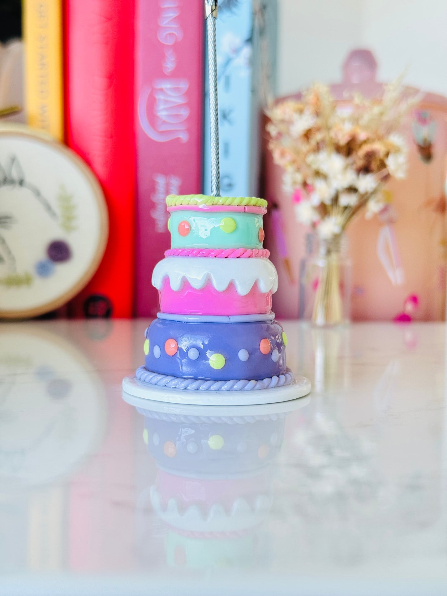 Colorful cake-shaped pen holder on a reflective surface with books and decorative items in the background.