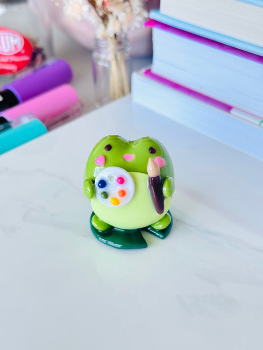 Green ceramic artist frog figurine with paintbrush and palette on a white surface
