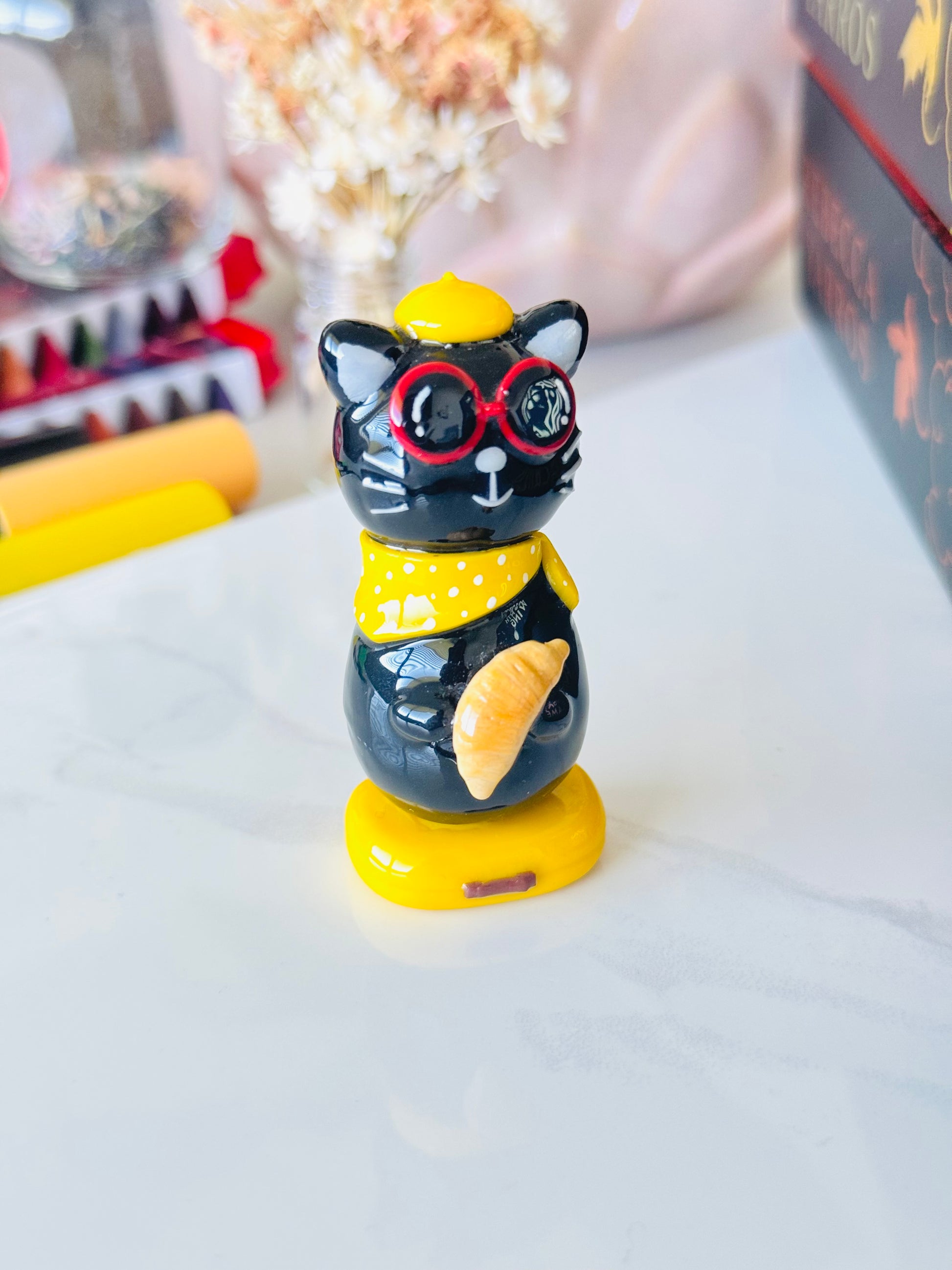 Black cat figurine with red glasses and yellow hat on a white surface
